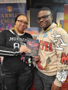 Smiling couple holding "Of Fear and Faith" by N.D. Jones at a book expo — readers celebrating fantasy that reflects them
